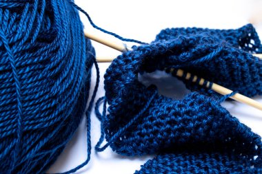 Midnight blue yarn, tangled and slightly astray next to beige knitting needles holding together a small classic pearl knit scarf. 