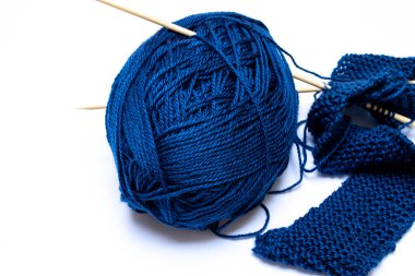 Midnight blue yarn, tangled and slightly astray next to beige knitting needles holding together a small classic pearl knit scarf. 