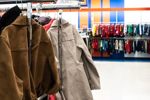 A suede jacket and a beige trench coat on a silver clothing rack in a thrift store in London, Ontario, Canada. Bokeh, soft-focus background showing kids apparel lined up on racks.