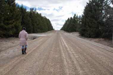 Pembe kabarık bir parka ve siyah bir toka ve kot pantolon giyen bir kadın, Mart 2021 'de, Ontario Canada' da kozalaklı ladin ağaçlarıyla kaplı kırsalda eski bir toprak yolda yürüyor..