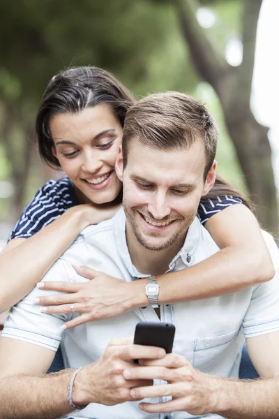 Casal feliz assistindo um telefone inteligente sentado em um banco em um parque — Fotografia de Stock
