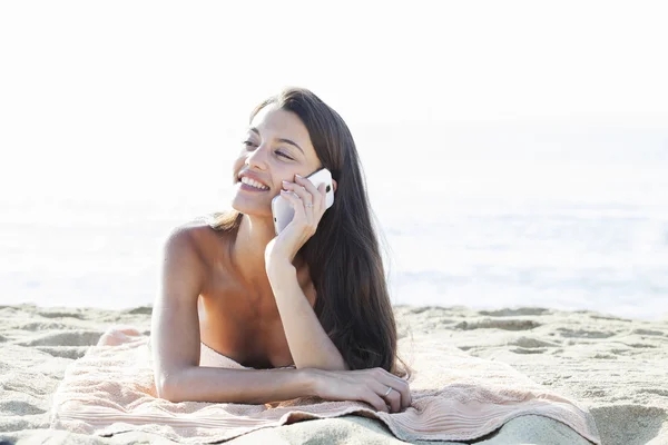 Portrait of an attractive girl laying down relaxing on holiday, using a smartphone device on vacation laying down on a beach towel on the sand — Stock Photo, Image