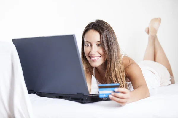 A smiling woman on her bed with laptop in front of her and credit card in hand