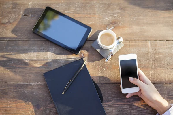 Cup of hot coffee, digital tablet, note book and woman hand using smartphone on wood table background