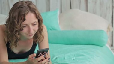 Smartphone ile yatakta yatan genç kadın kıvırcık. Dolly vurdu