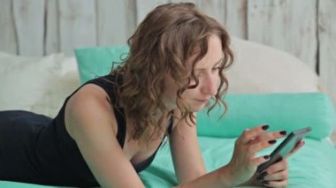 Smartphone ile yatakta yatan genç kadın kıvırcık. Dolly vurdu