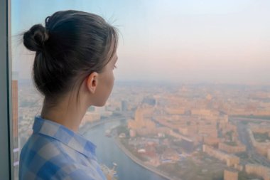 Düşünceli bir kadın gökdelenin arka penceresinden şehir manzarasına bakıyor.