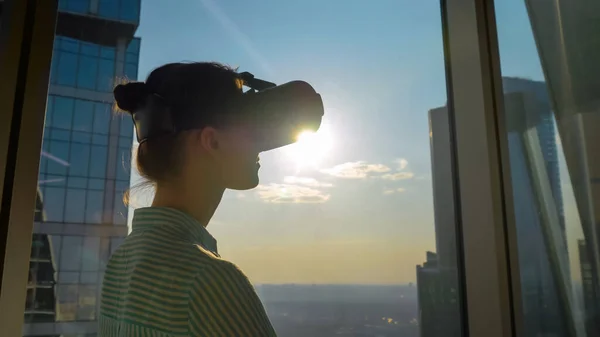 Young woman using virtual reality headset against skyscraper window - VR concept