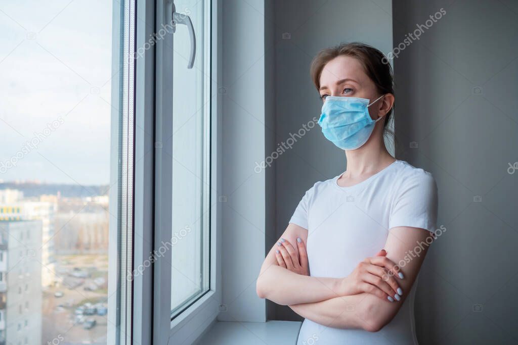 Pensive woman wearing medical face mask and looking out of window at home