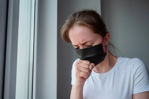 Woman with black face mask suffering from coughing, looking out of window