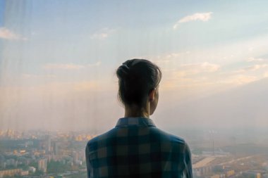 Düşünceli bir kadın gökdelenin arka penceresinden şehir manzarasına bakıyor.