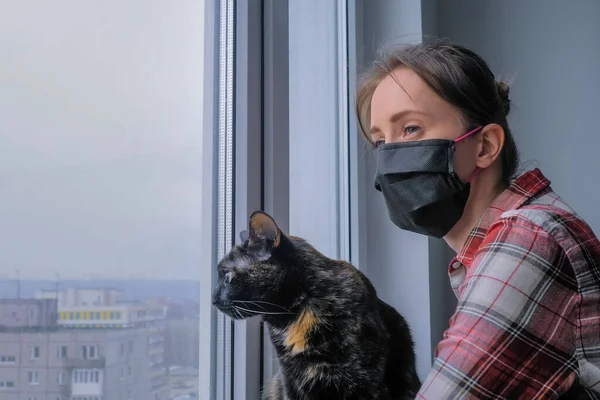 Black cat and woman in black medical face mask looking out of window at home