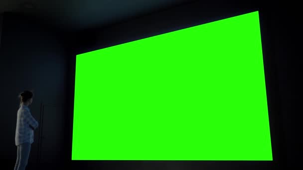 Kobieta patrząc na duży pusty zielony ekran w ciemnym pokoju - koncepcja klucza chroma — Wideo stockowe