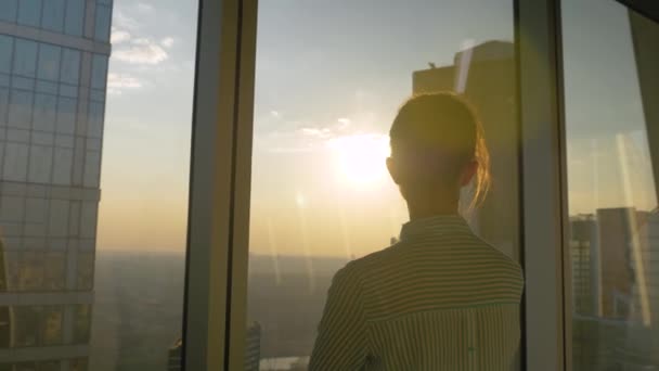 Vue de dos : femme regardant le paysage urbain à travers la fenêtre du gratte-ciel au coucher du soleil — Video