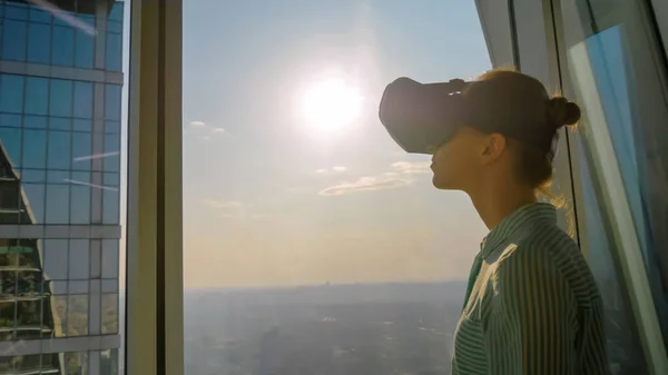 Young woman using virtual reality headset against skyscraper window - VR concept