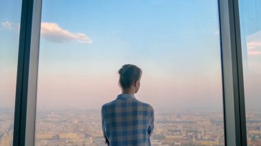 Düşünceli bir kadın gökdelenin arka penceresinden şehir manzarasına bakıyor.