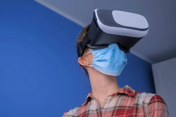 Young woman wearing medical face mask, virtual reality headset against blue wall