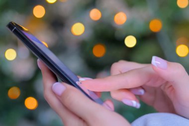 Akıllı telefon kullanan bir kadın, Noel ışığı arka planı - yan görünümü kapat