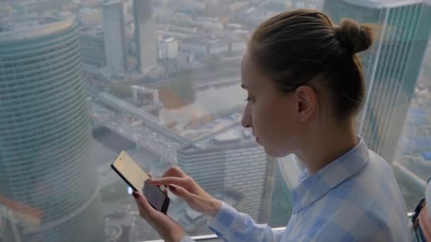 Mujer usando smartphone contra vista de paisaje urbano a través de ventana de rascacielos — Vídeos de Stock