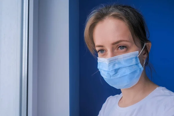 Pensive woman wearing medical face mask and looking out of window - close up