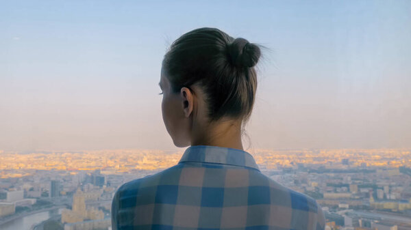 Back view of pensive woman looking at cityscape through window of skyscraper