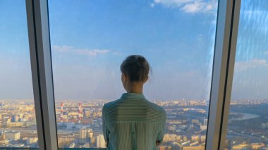 Düşünceli bir kadın gökdelenin arka penceresinden şehir manzarasına bakıyor.