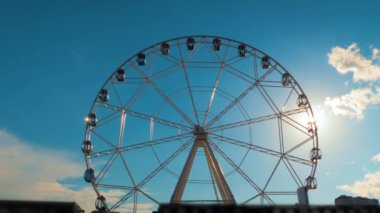 Dönme dolabın silueti bulutlarla mavi gökyüzüne doğru dönüyor: güneş merceği parlıyor