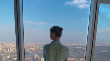 Düşünceli bir kadın gökdelenin arka penceresinden şehir manzarasına bakıyor.