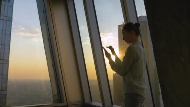 Retrato de mujer usando smartphone contra el paisaje urbano al atardecer a través de la ventana — Vídeos de Stock