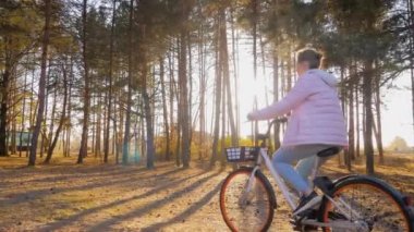 Sonbahar parkında gün batımında bisiklete binen genç bir kadın. Ağır çekim.