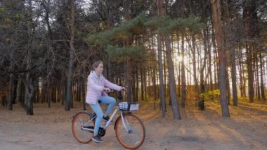 Yavaş çekim: Sonbahar şehir parkında gün batımında bisiklet süren genç bir kadın.