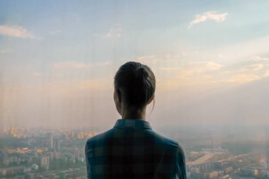 Düşünceli bir kadın gökdelenin arka penceresinden şehir manzarasına bakıyor.