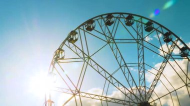 Dönme dolabın silueti bulutlarla mavi gökyüzüne doğru dönüyor: güneş merceği parlıyor