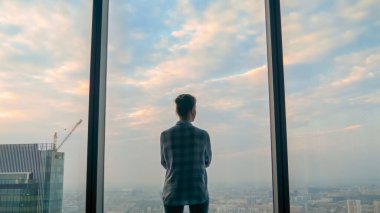 Düşünceli bir kadın gökdelenin arka penceresinden şehir manzarasına bakıyor.
