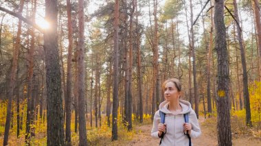 Sırt çantalı bir kadın sonbahar parkında yürüyor, güneş gözlüğü parlıyor.