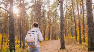 Sırt çantalı bir kadın sonbahar parkında yürüyor, güneş gözlüğü parlıyor.