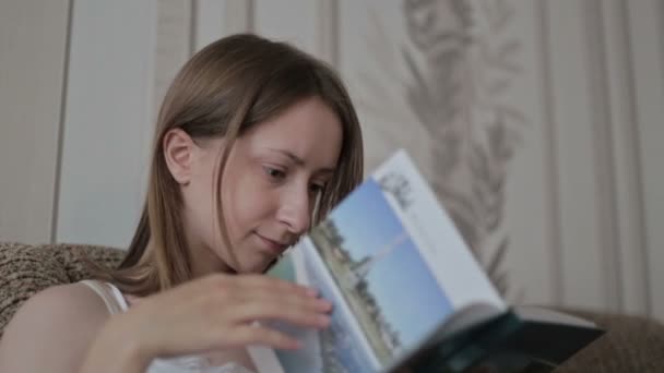 Fiatal nő olvas egy könyvet a kanapén — Stock videók