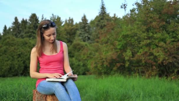 Молодая женщина сидит в парке и читает книгу — стоковое видео