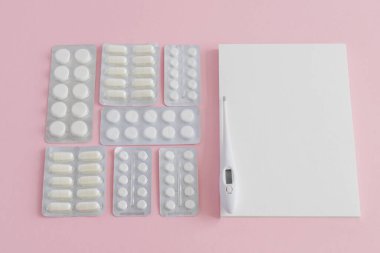 Tıbbi ilaçlar, haplar, vücut ısısını ölçmek için beyaz bir termometre ve pembe arka plan üzerine notlar için boş bir defter..