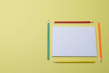 Sarı bir arka planda, renkli kalemler ve beyaz boş bir kâğıt sayfasında, yazı için bir yer, yatay fotoğraf üst görünümü.