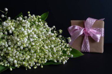 Bir buket kokulu beyaz vadi zambakları koyu bir arka planda, pakette pembe kurdeleli bir hediyeyle. Stüdyo fotoğrafı. Tebrik kartı. Yatay fotoğraf.