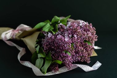 Tebrik konsepti, hediye. Bir demet mor leylak, esnaf kağıdına sarılmış koyu arkaplanda pembe saten kurdeleler..