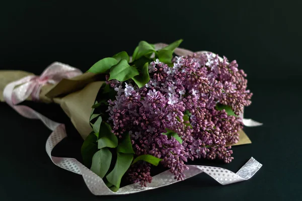 Tebrik konsepti, hediye. Bir demet mor leylak, esnaf kağıdına sarılmış koyu arkaplanda pembe saten kurdeleler..