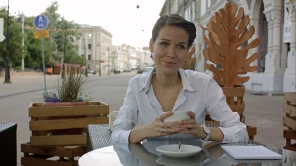 Mujer tomando un café de una taza en la terraza de un restaurante mientras piensa y mira hacia los lados — Vídeos de Stock