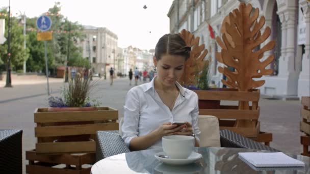 Junge Frau trinkt Kaffee und benutzt ihr Handy in einem Café im Freien — Stockvideo
