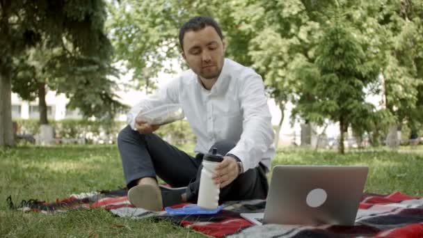 Ung affärsman sitter i gräset och äter lunch i en park på sommaren. — Stockvideo