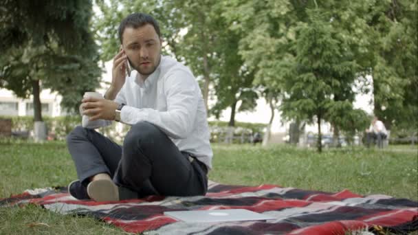 İstediğin zaman ara. Parkta çimlere oturup telefonda konuşan neşeli genç adam — Stok video