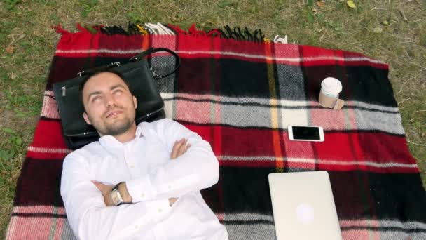 Uomo d'affari moderno e rilassante con le mani dietro la testa sull'erba con computer portatile nelle vicinanze — Video Stock