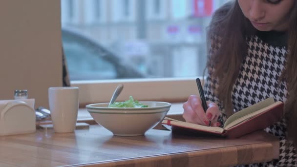 Paže mladé ženy, píše v knize poznámek. Sedí u stolu v kavárně a žere salát — Stock video