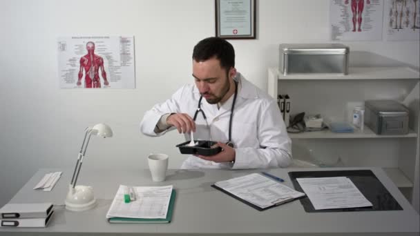 Médecin barbu étouffé lors d'un déjeuner à son bureau. Il mange dans un récipient en plastique, boit dans une tasse et pense à quelque chose. — Video